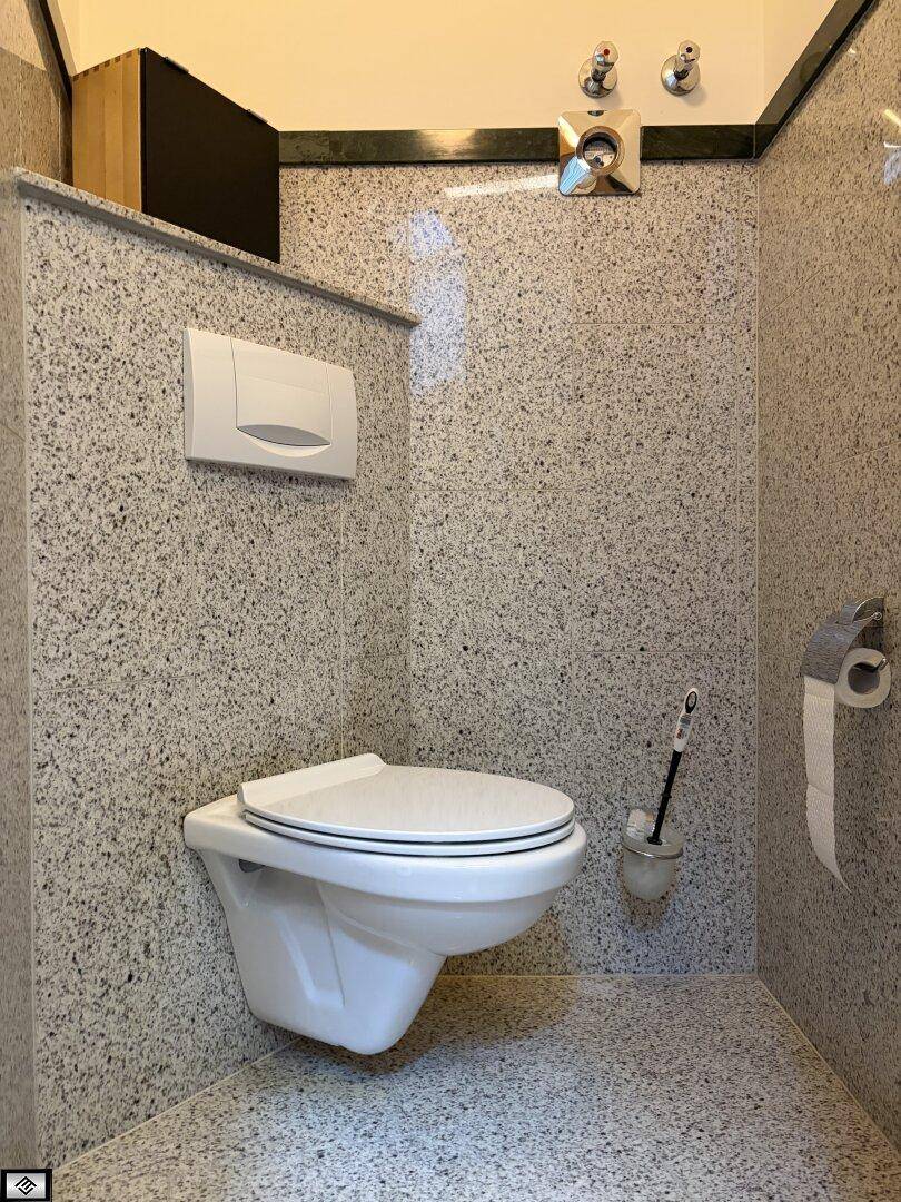 Separates WC mit wandhängender Toilette und gefliesten Wänden in hellem Granit-Look.