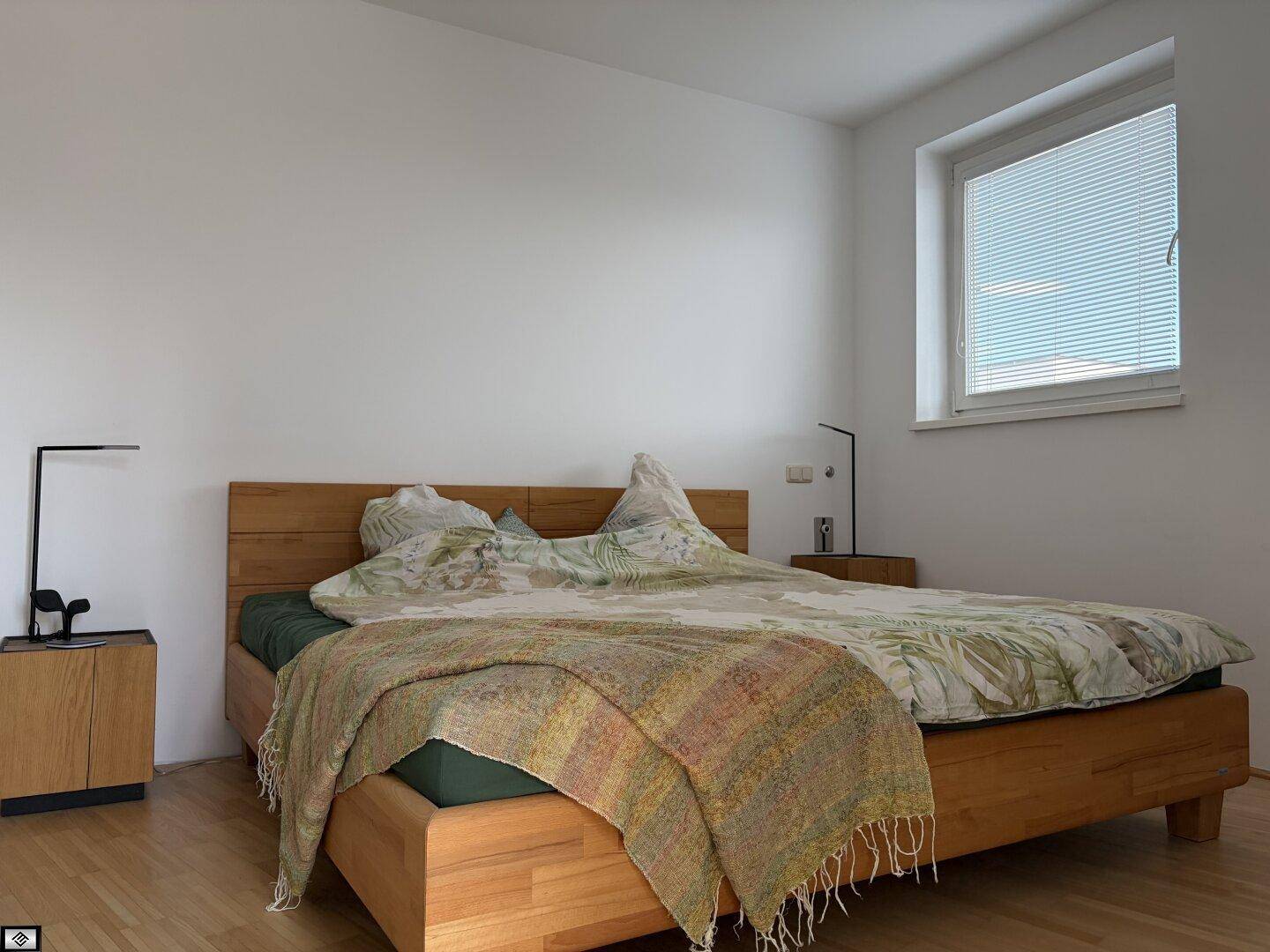 Helles Schlafzimmer mit einem Holzbett, Nachttischen und einem Fenster mit Jalousien.