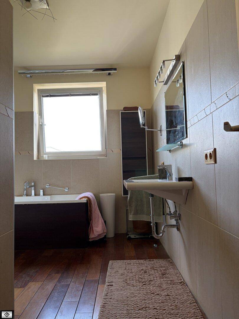 Badezimmer mit Badewanne, Waschbecken, Spiegel und Fenster, ausgestattet mit dunklem Holzboden.