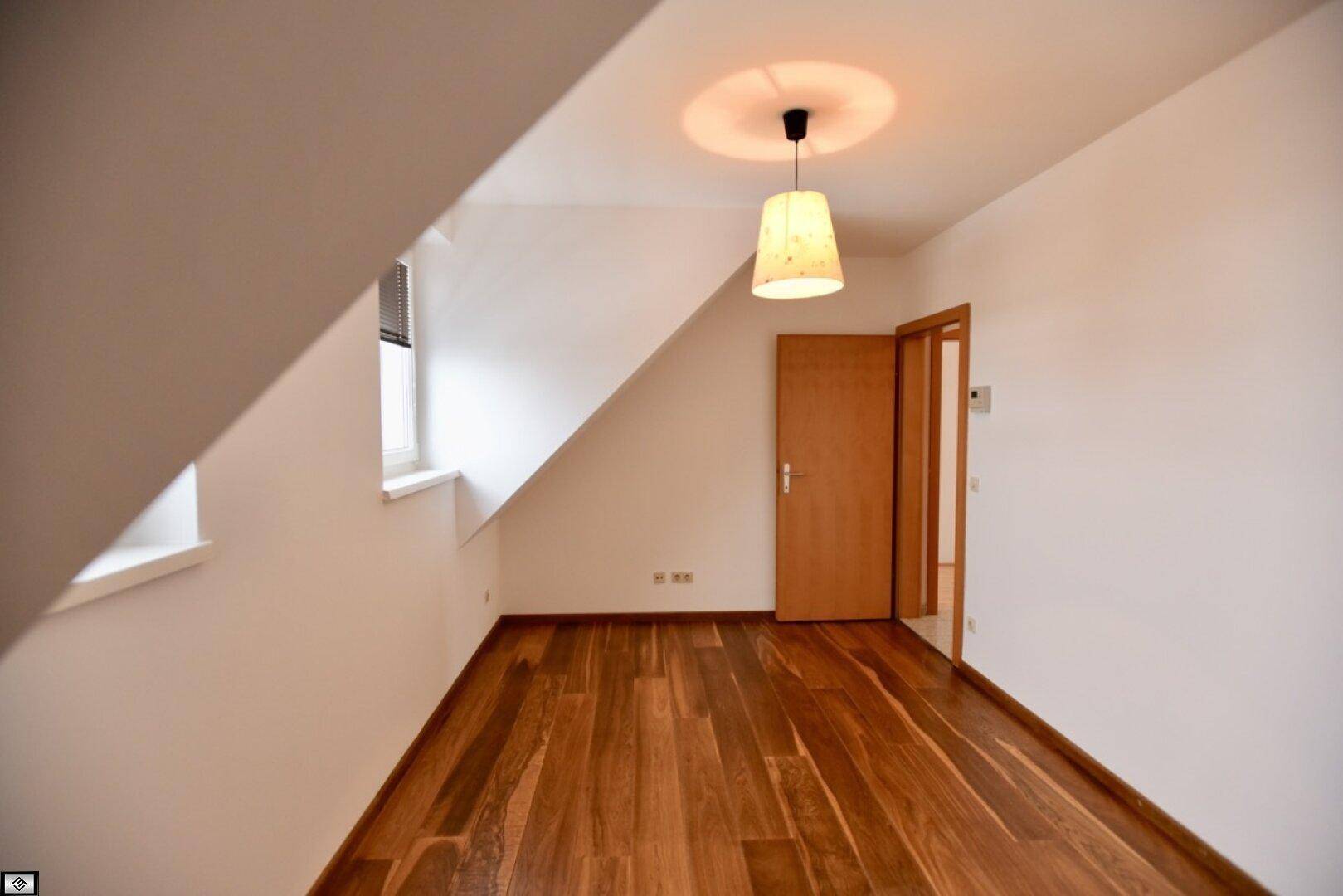 Leerer Raum mit Dachschräge, Fenster und dunklem Holzplankenboden, bereit zur Einrichtung.