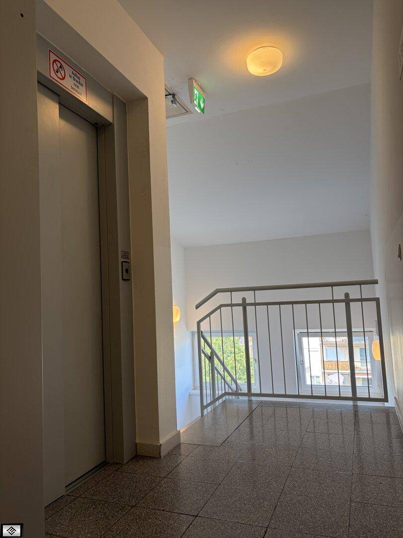 Heller Flur mit Aufzug und Treppenhaus, ausgestattet mit Fliesenboden und Metallgeländer.