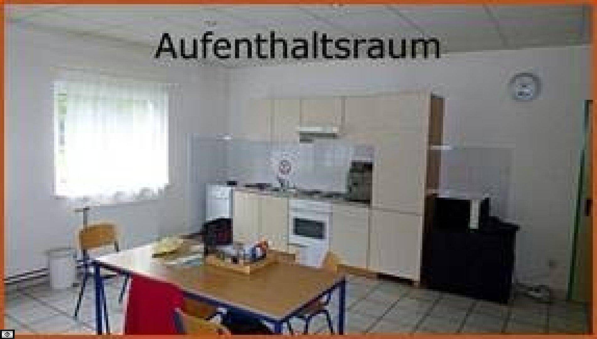 Aufenthaltsraum mit kleiner Küchenzeile, Esstisch und Stühlen, sowie einem Fenster mit Vorhang.