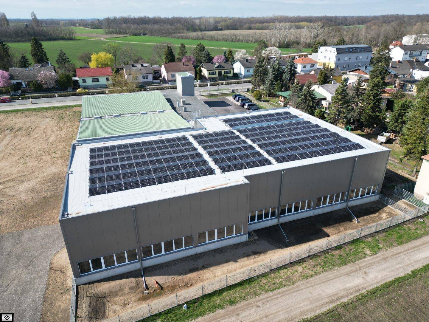 Drohnenansicht eines großen Industriegebäudes mit Photovoltaikanlage auf dem Dach und angrenzender Wohnbebauung.