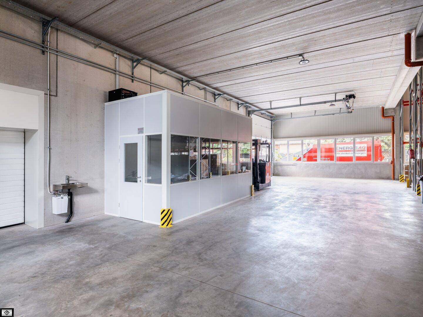 Geräumige Lagerhalle mit Betonboden, einer integrierten Bürobox und sichtbaren Installationen an der Decke.