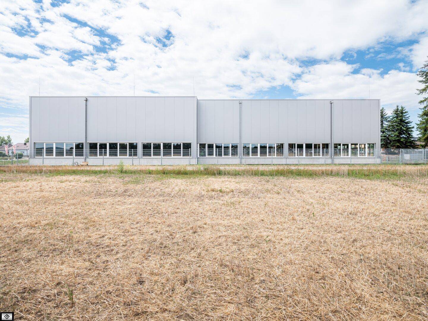 Lange Fassade eines modernen Industriegebäudes mit vielen Fenstern und einem weiten Feld im Vordergrund.