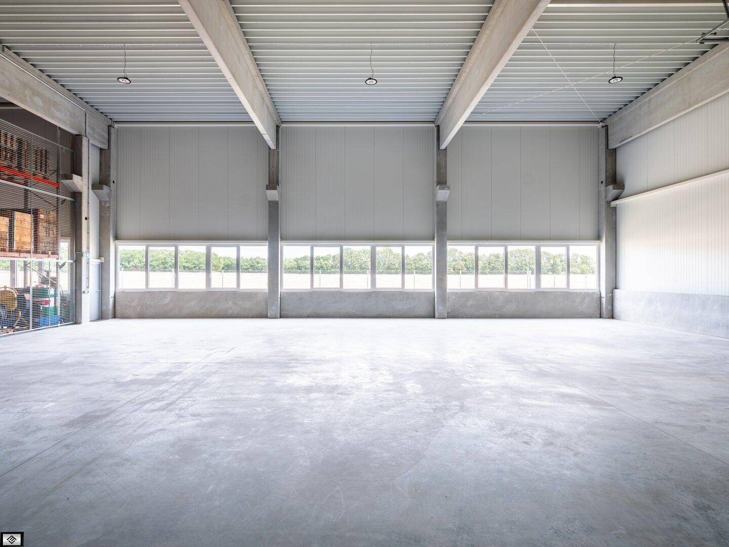 Weitläufige, leere Lagerhalle mit hohen Decken, Betonboden und einer Reihe von Fenstern für Tageslicht.