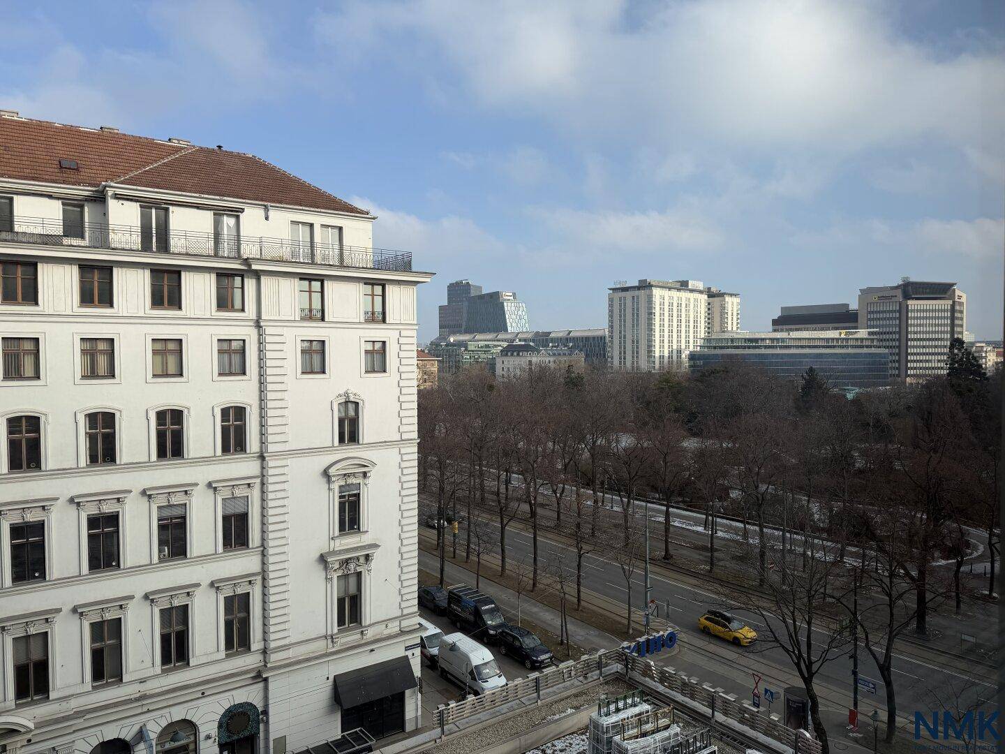 Blick von oben auf eine belebte Stadtstraße mit Gebäuden und Bäumen unter blauem Himmel.