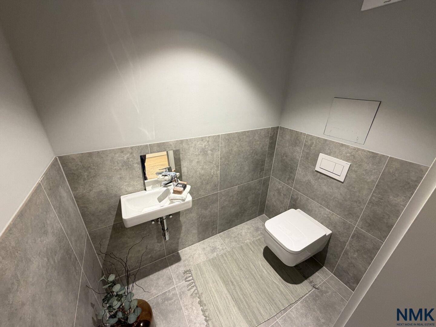 Modernes Gäste-WC mit grauen Fliesen, wandhängendem WC und kleinem Waschbecken.