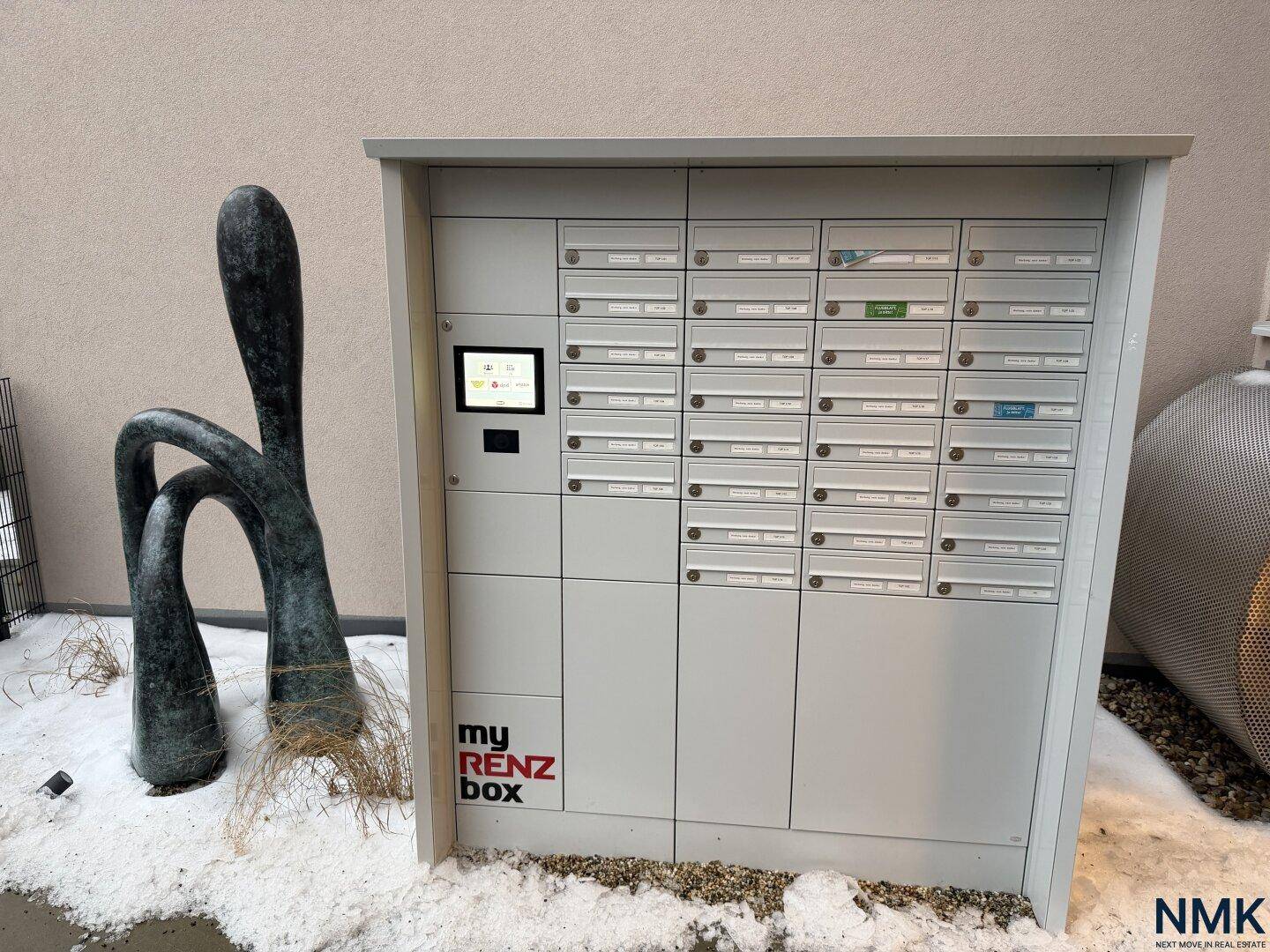 Modernes Briefkasten- und Paketboxensystem im Außenbereich, umgeben von Schnee und einer Skulptur.