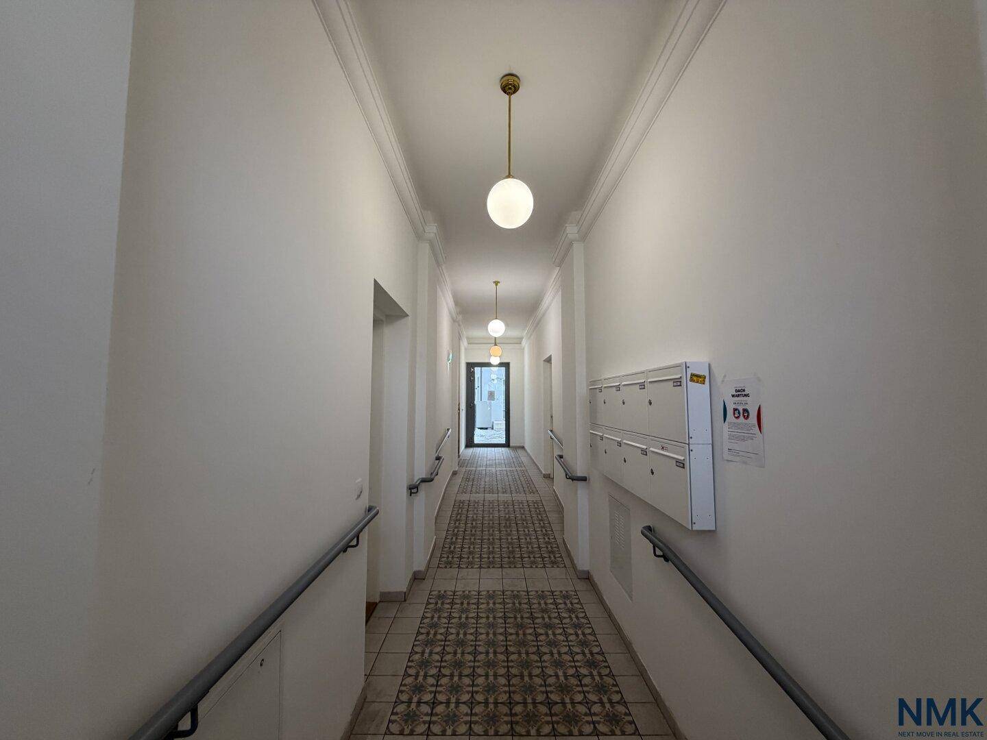 Langer, heller Hausflur mit Fliesenboden, Handläufen und Briefkästen.