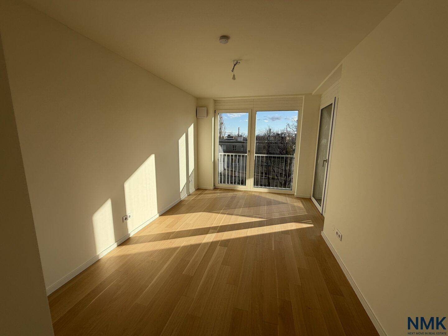 Unmöbliertes Schlafzimmer mit Holzboden und großen Fenstern, die Zugang zu einem Balkon bieten.