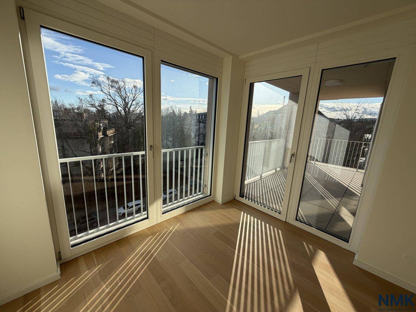 Heller Raum mit Holzboden und großen Fenstern, die viel Sonnenlicht hereinlassen und Zugang zum Balkon bieten.