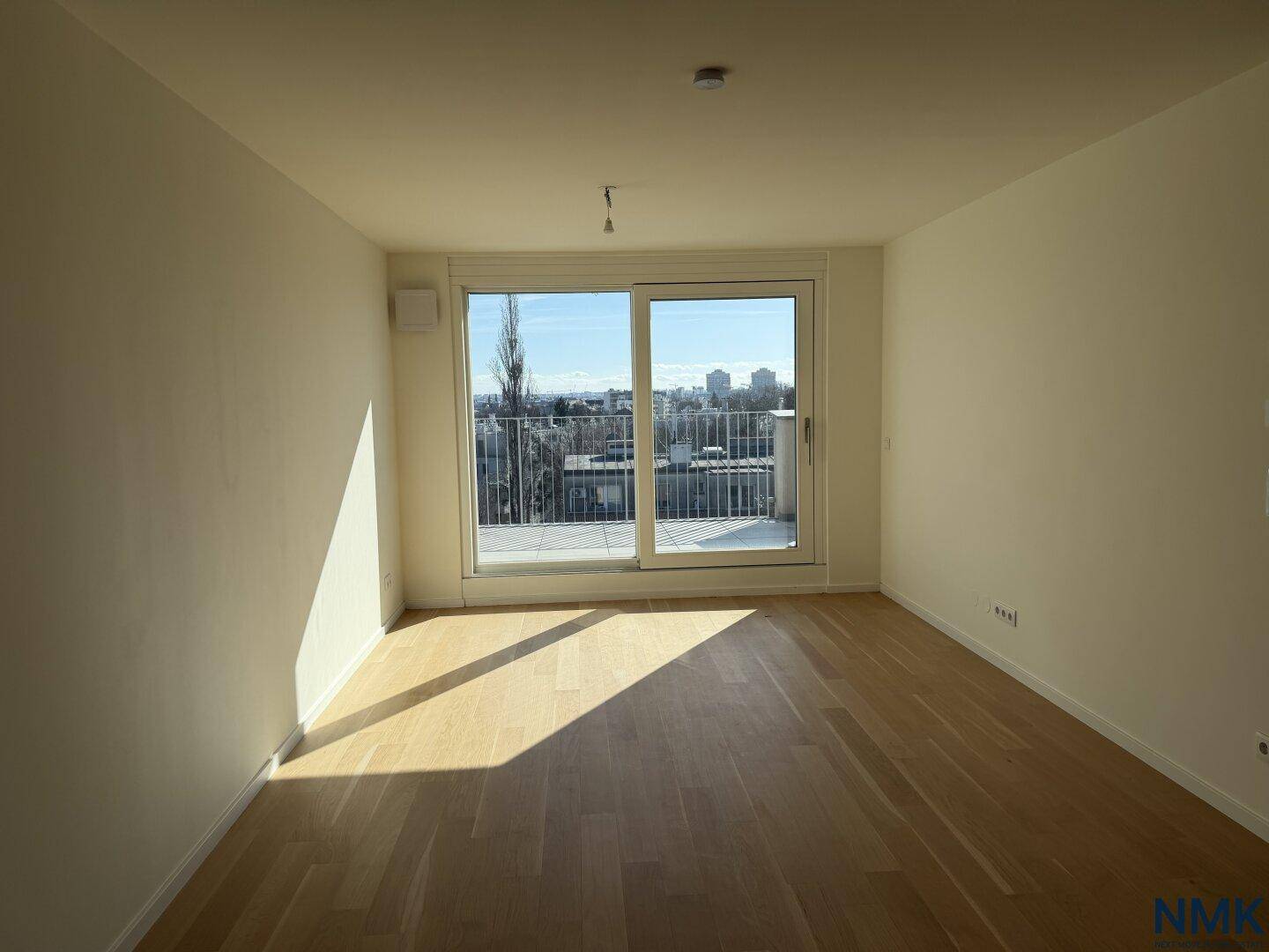 Helles, leeres Wohnzimmer mit Holzboden und großen Fenstern, die auf einen Balkon führen.