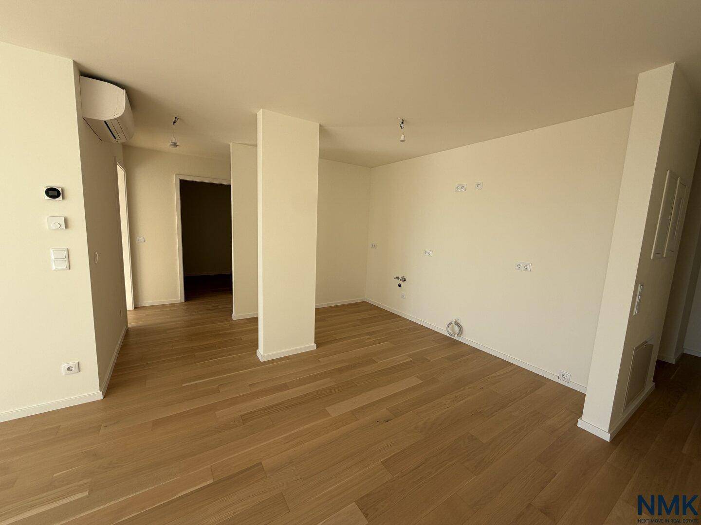 Leerer Küchenbereich mit Holzboden, Klimaanlage und vorbereiteten Anschlüssen für eine Einbauküche.