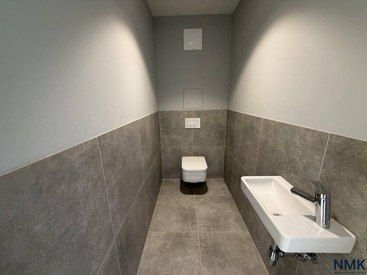 Modernes Gäste-WC mit grauen Fliesen, wandhängendem WC und kleinem Waschbecken.