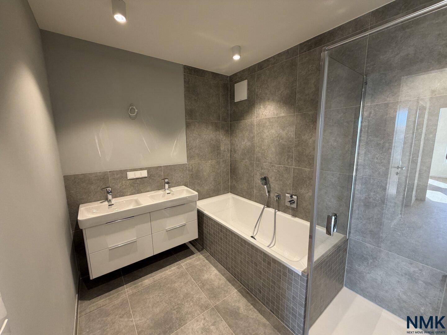 Modernes Badezimmer mit grauen Fliesen, Badewanne, Dusche und Doppelwaschbecken.
