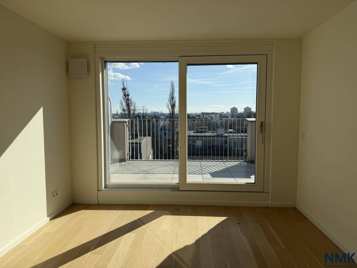 Leeres Zimmer mit Holzboden und großen Schiebetüren, die zu einer Terrasse mit Stadtblick führen.