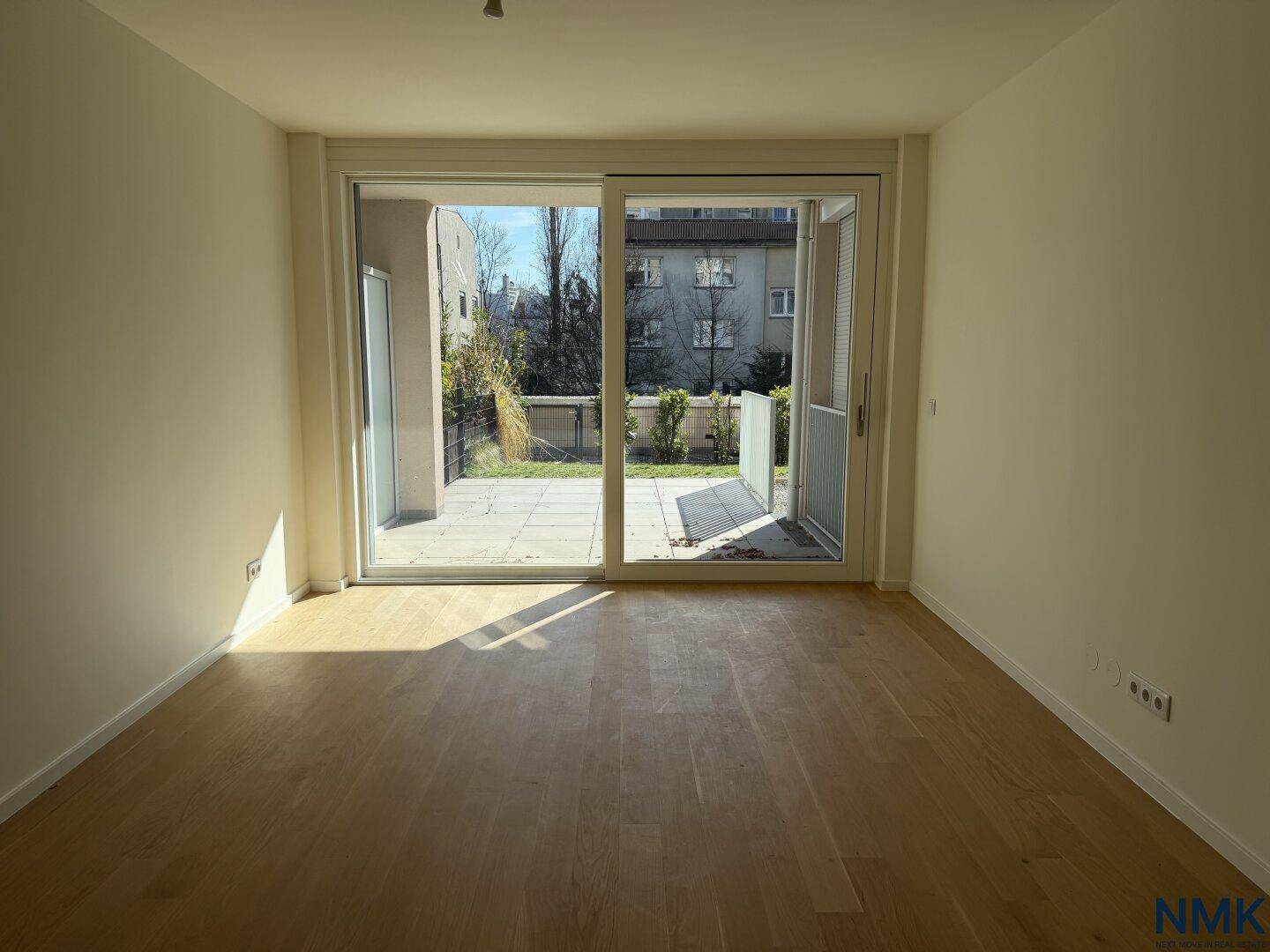 Geräumiges, leeres Wohnzimmer mit Holzboden und direktem Zugang zu einer sonnigen Terrasse.