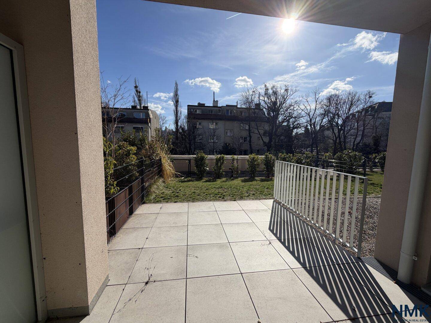 Sonnige Terrasse mit Fliesenboden und Blick auf den Gemeinschaftsgarten und umliegende Gebäude.