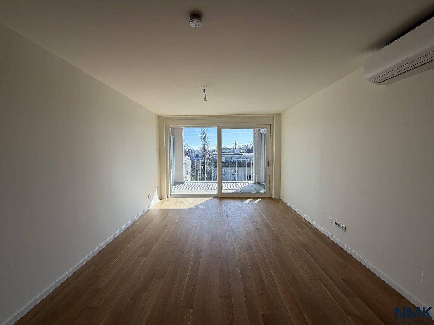 Geräumiges, leeres Wohnzimmer mit Holzboden und großem Balkonzugang.