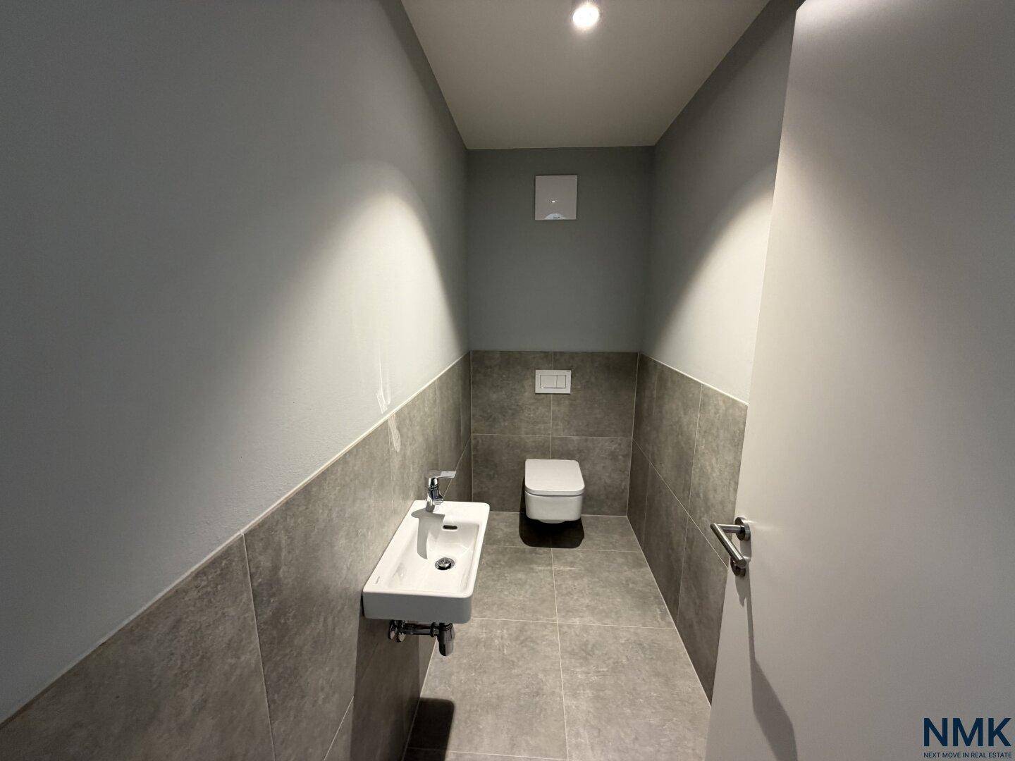 Modernes Gäste-WC mit grauen Fliesen, wandhängendem WC und kleinem Waschbecken.