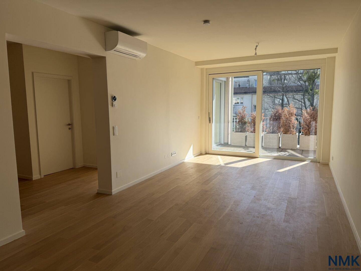 Geräumiges, leeres Wohnzimmer mit Holzboden, Klimaanlage und großem Balkonzugang.