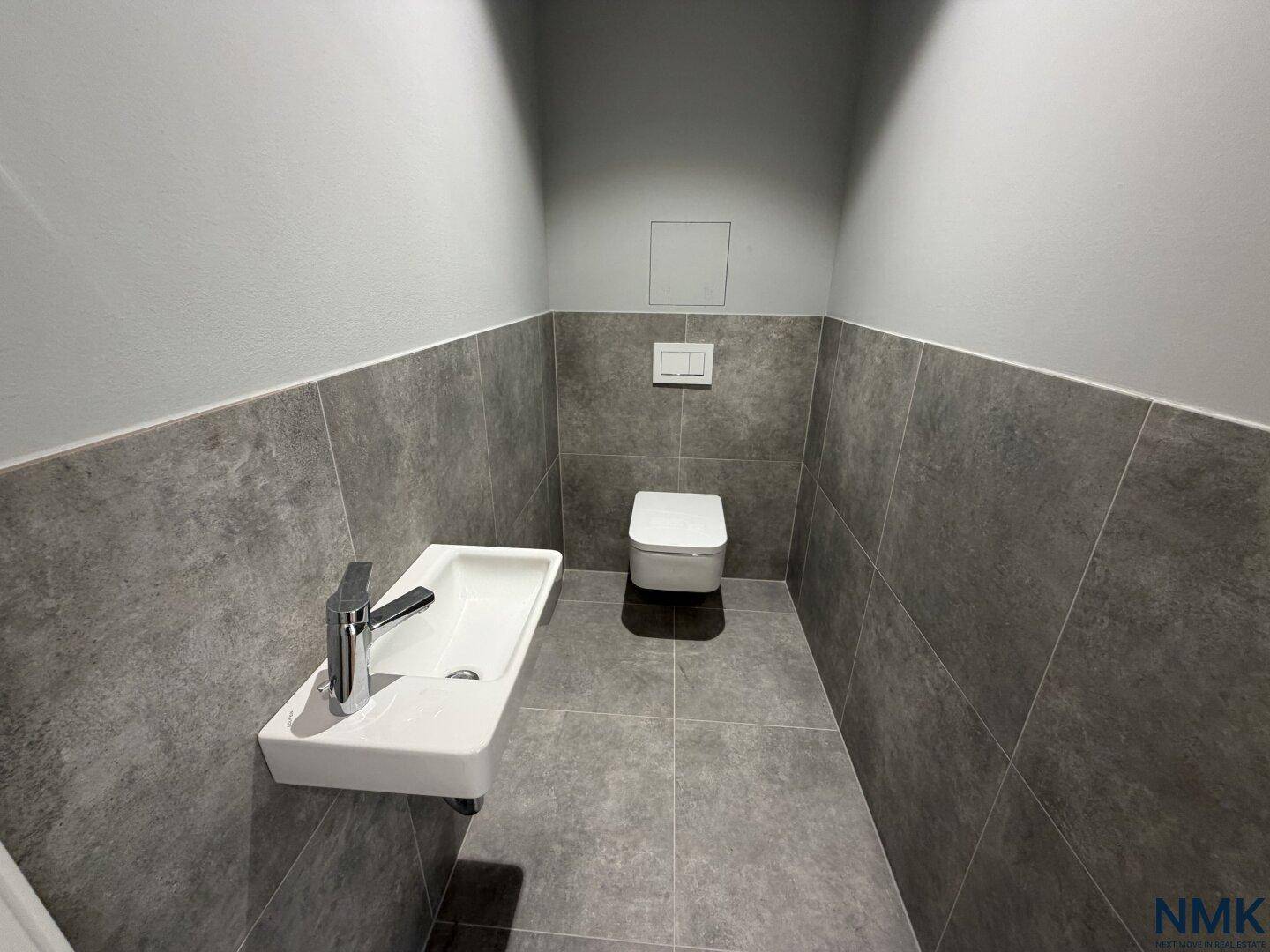 Modernes Gäste-WC mit grauen Fliesen, wandhängendem WC und kleinem Waschbecken.