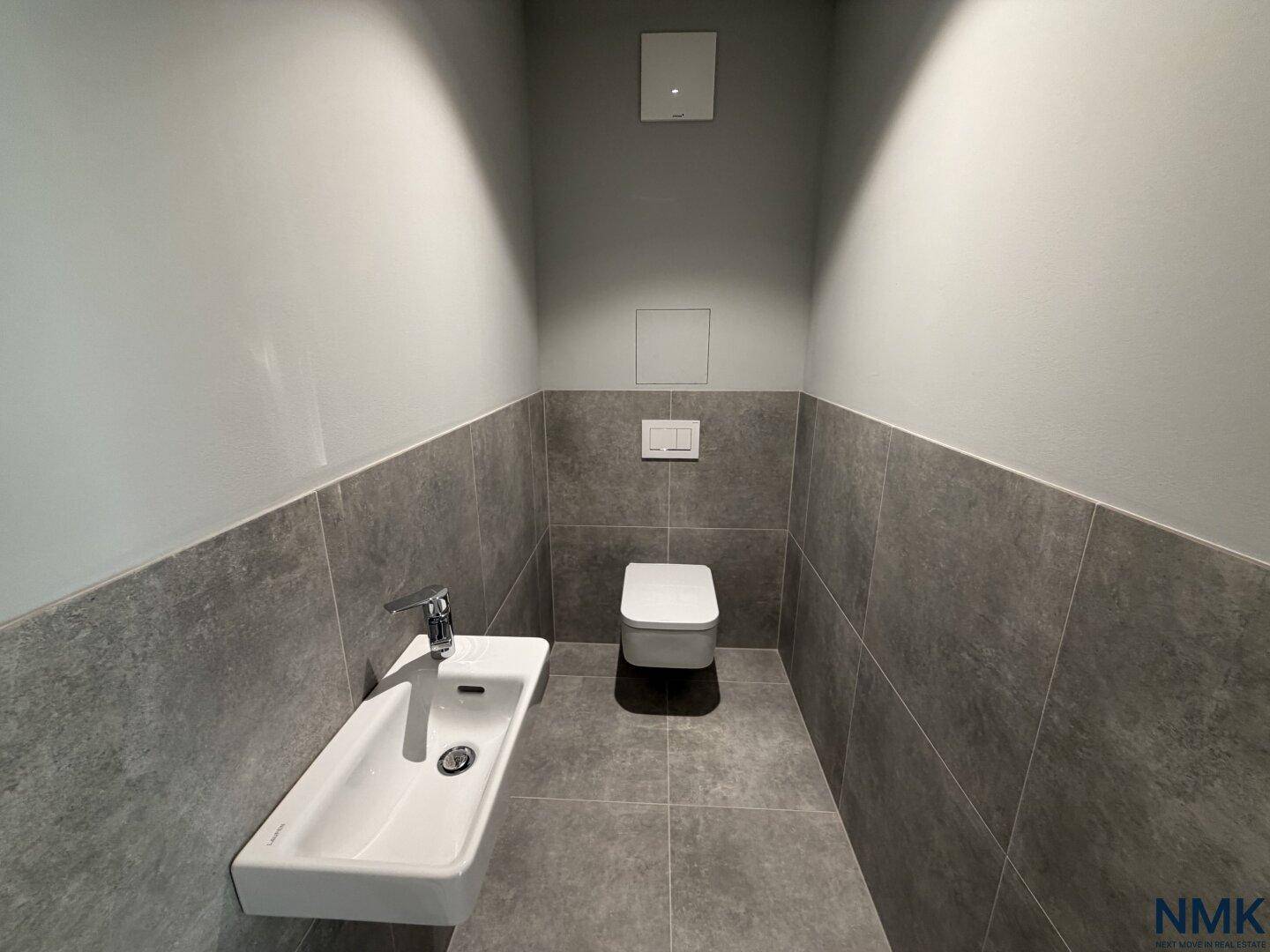 Modernes Gäste-WC mit grauen Fliesen, wandhängendem WC und kleinem Waschbecken.