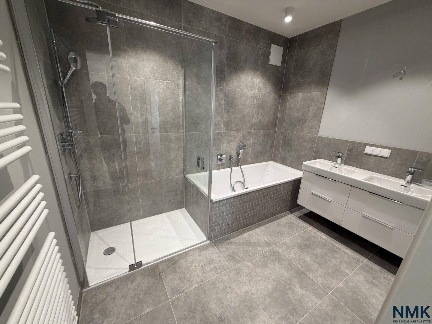 Modernes Badezimmer mit grauen Fliesen, Badewanne, Dusche und Doppelwaschbecken.