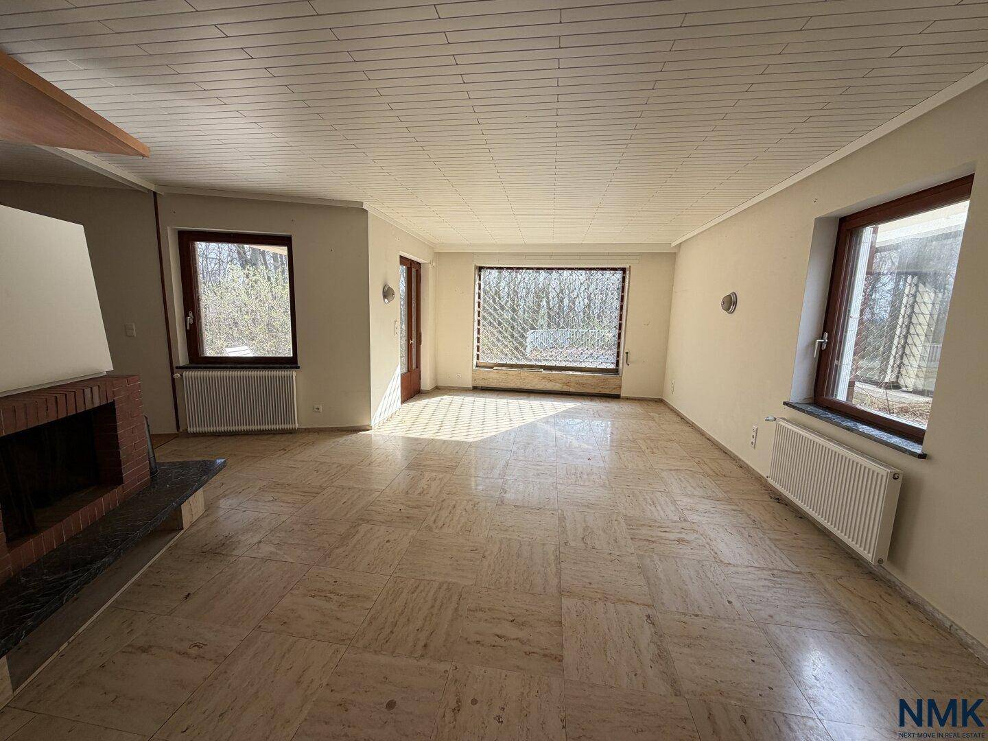 Leeres Wohnzimmer mit Fliesenboden, Kamin und großen Fenstern mit Blick ins Grüne.