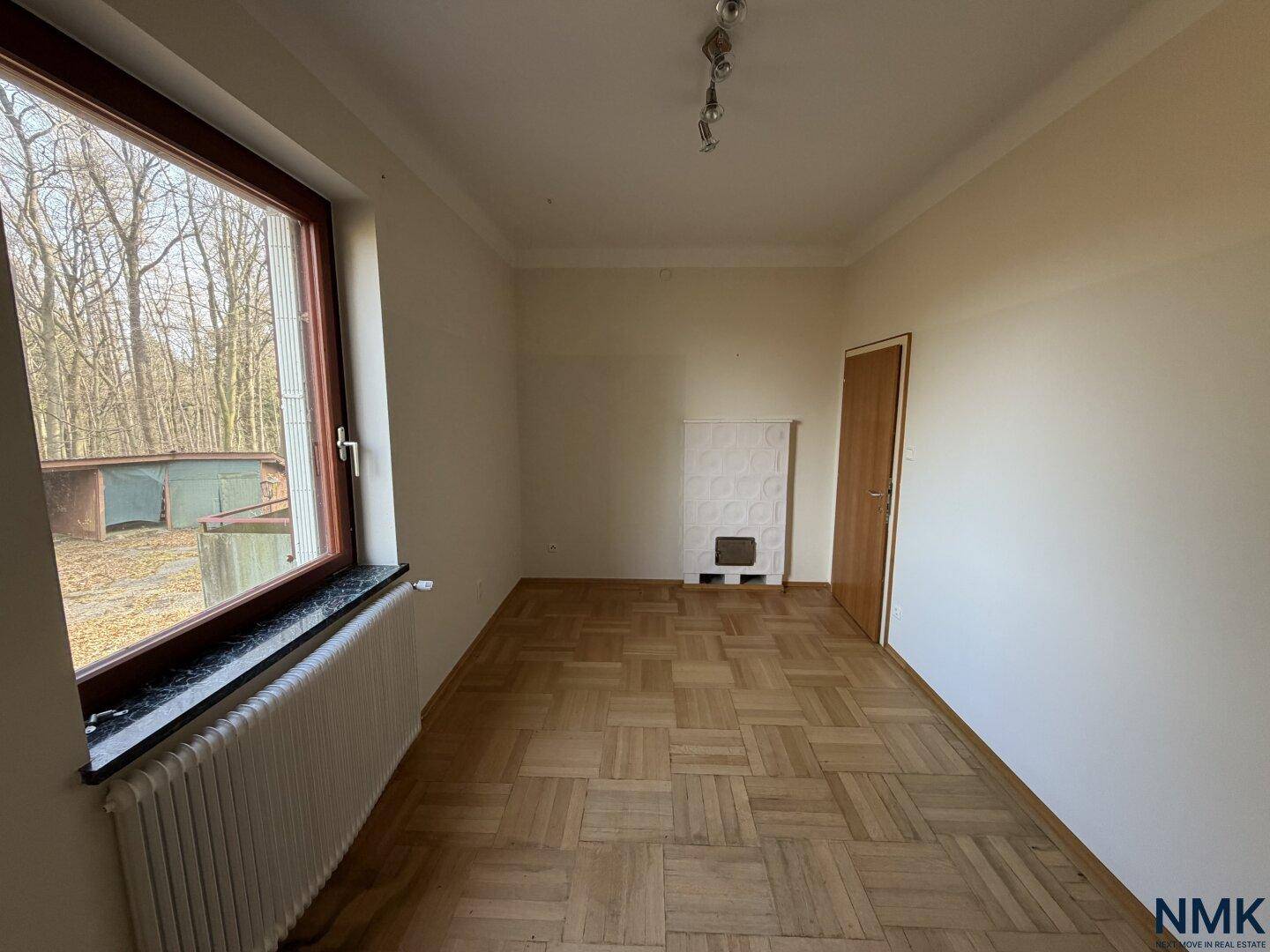 Leerer Raum mit Parkettboden und einem traditionellen Kachelofen, hell durch ein Fenster.