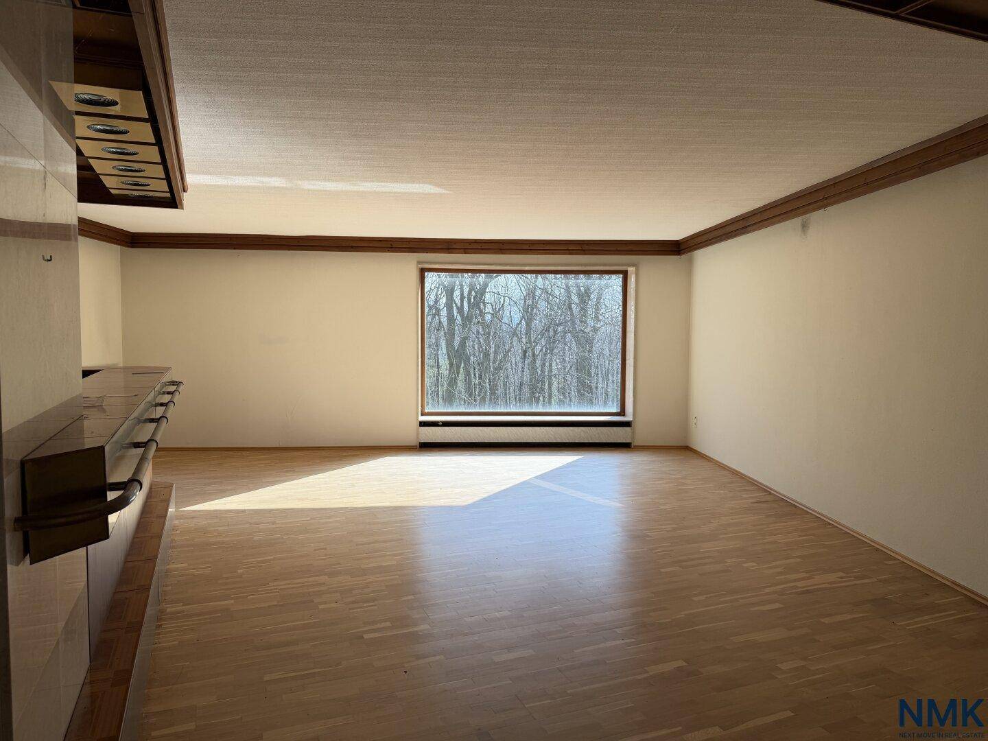 Großer leerer Raum mit Parkettboden, Barbereich und einem großen Fenster mit Waldblick.