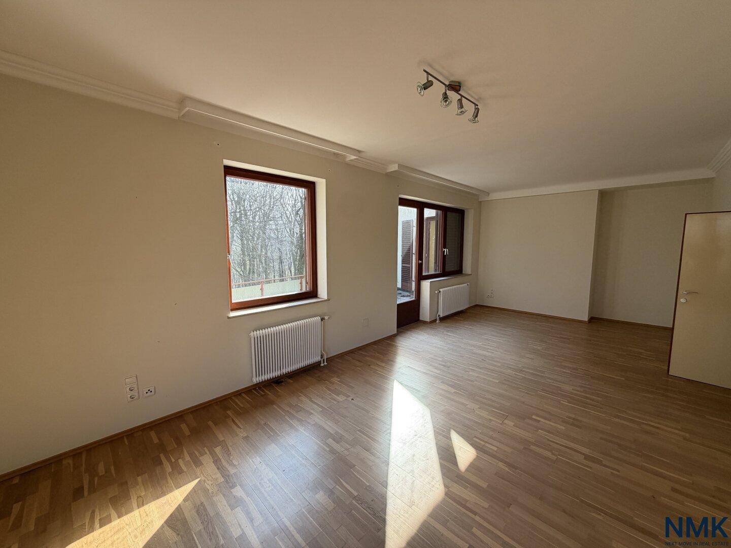 Heller, leerer Raum mit Holzfußboden, großem Fenster und Zugang zu einem Außenbereich.