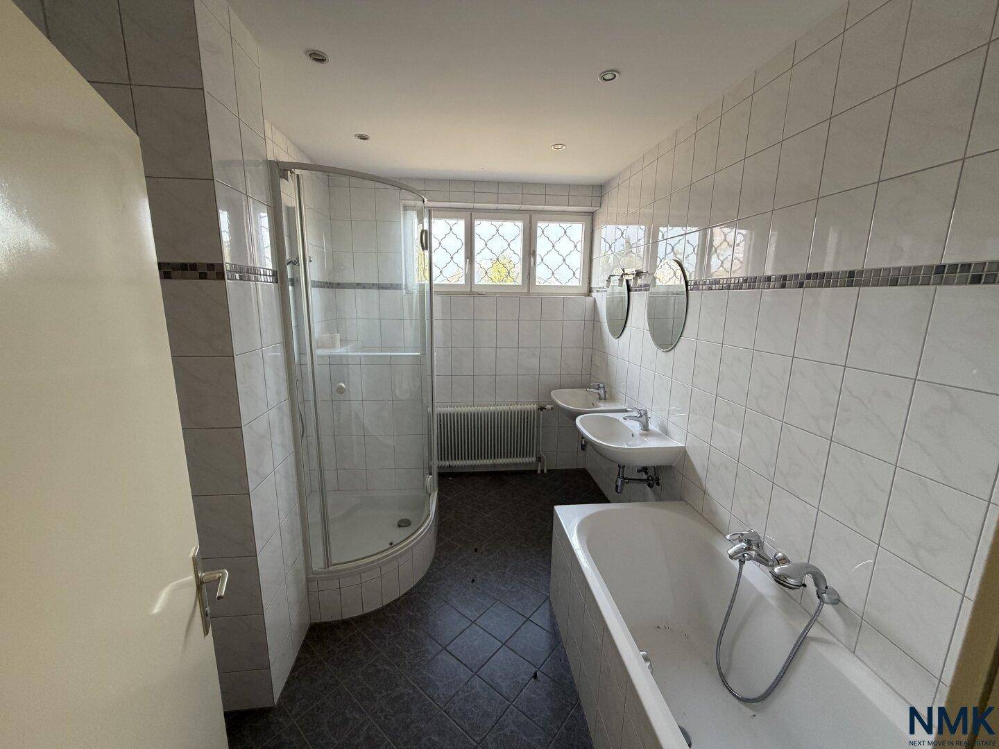 Gefliestes Badezimmer mit Duschkabine, Badewanne, Doppelwaschbecken und Fenster.