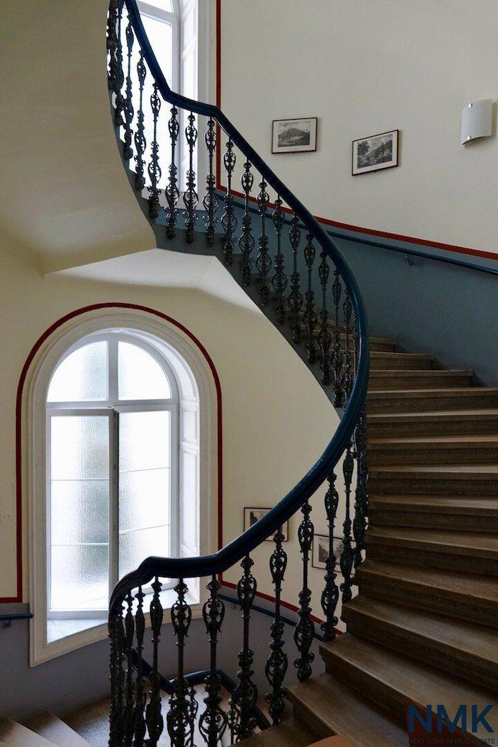 Elegantes Treppenhaus mit geschwungener Balustrade und hohen Bogenfenstern.