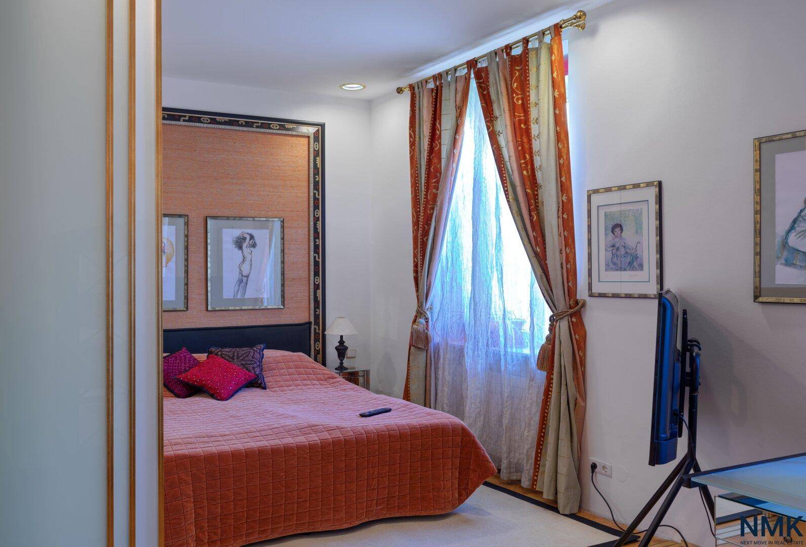 Gemütliches Schlafzimmer mit orangefarbener Bettdecke, Vorhängen und einem Fernseher.