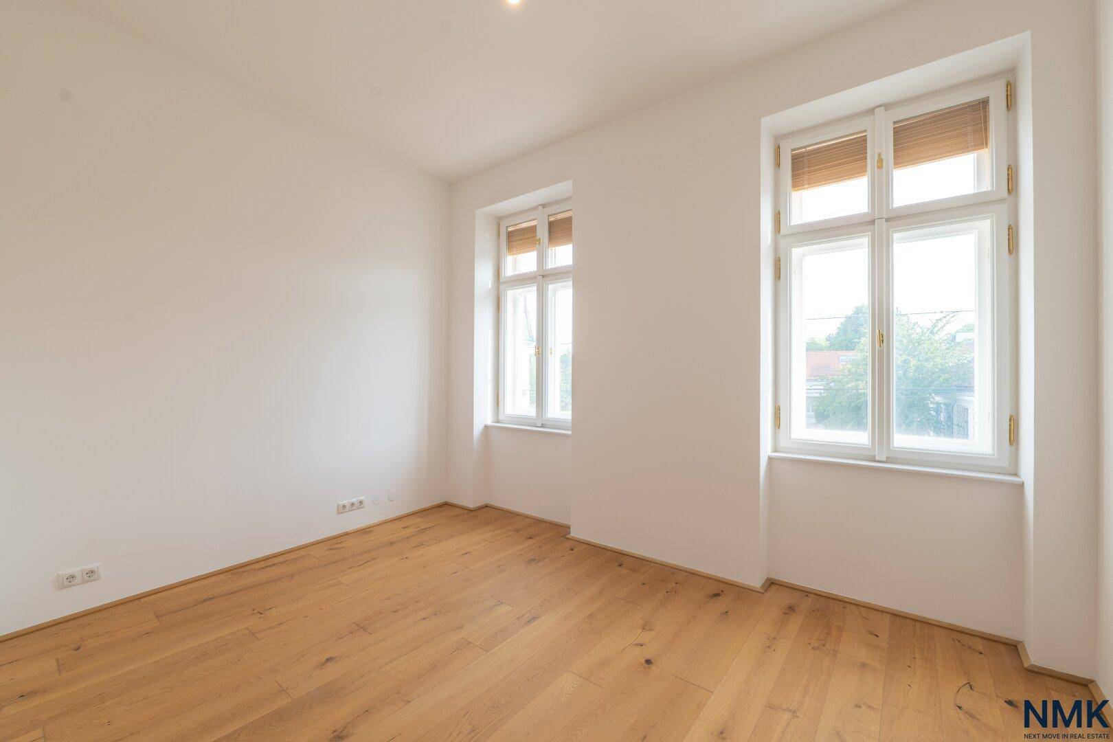 Geräumiges Zimmer mit Holzboden und großen Fenstern, die viel Tageslicht hereinlassen.