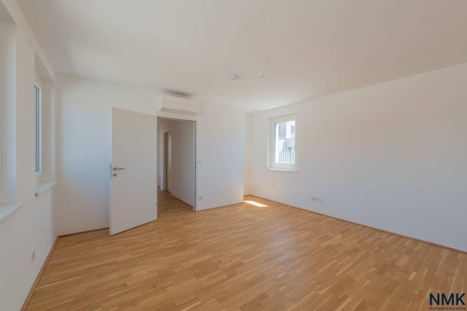 Geräumiges, leeres Schlafzimmer mit hellem Parkettboden, weißen Wänden und mehreren Fenstern.