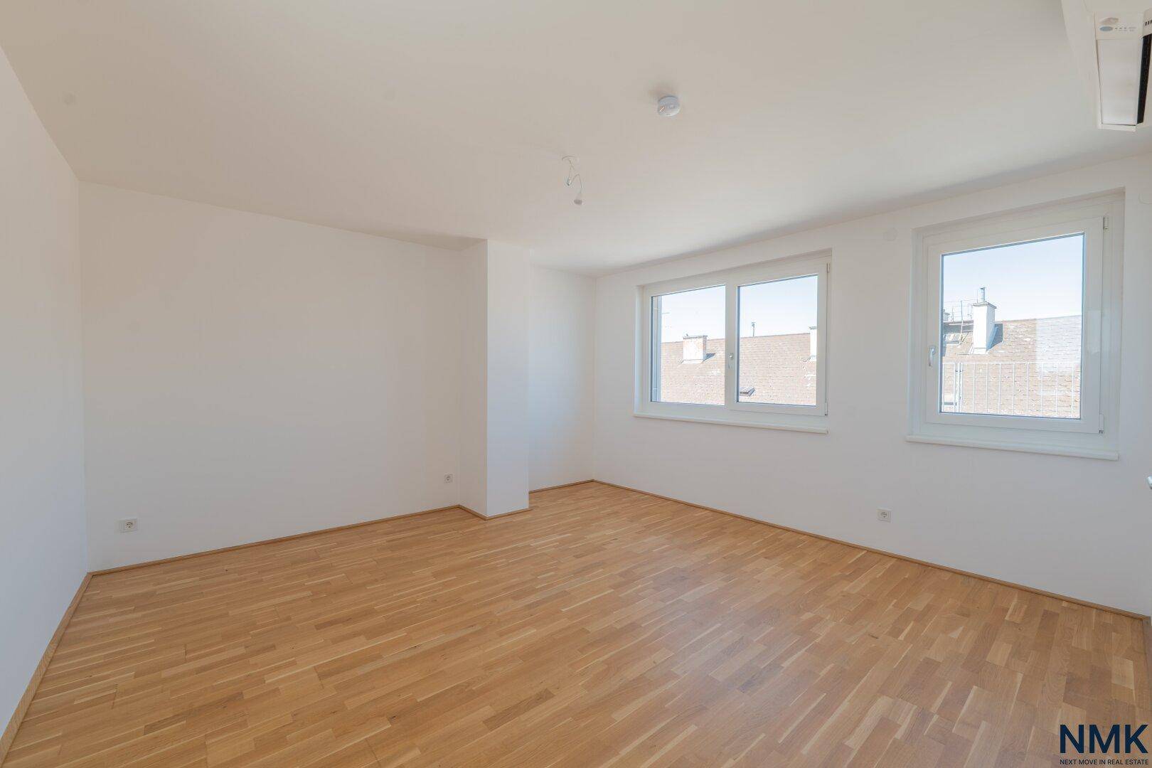 Helles, unmöbliertes Zimmer mit Parkettboden, weißen Wänden und zwei großen Fenstern.