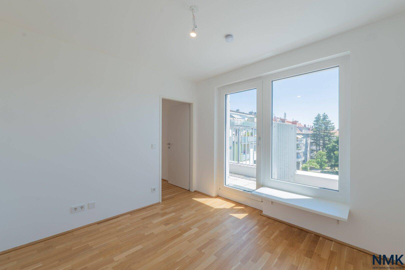 Heller, leerer Raum mit Parkettboden und großem Fenster mit Zugang zum Balkon.