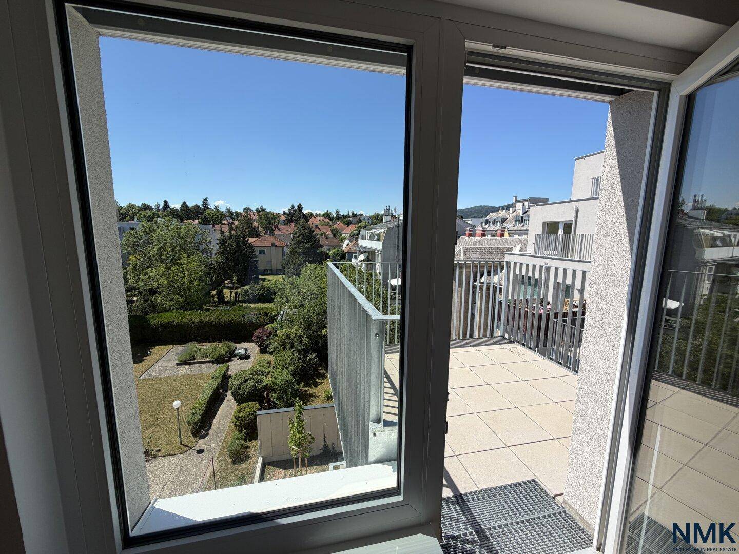Blick aus einem hellen Raum auf den Balkon und die grüne Umgebung unter blauem Himmel.