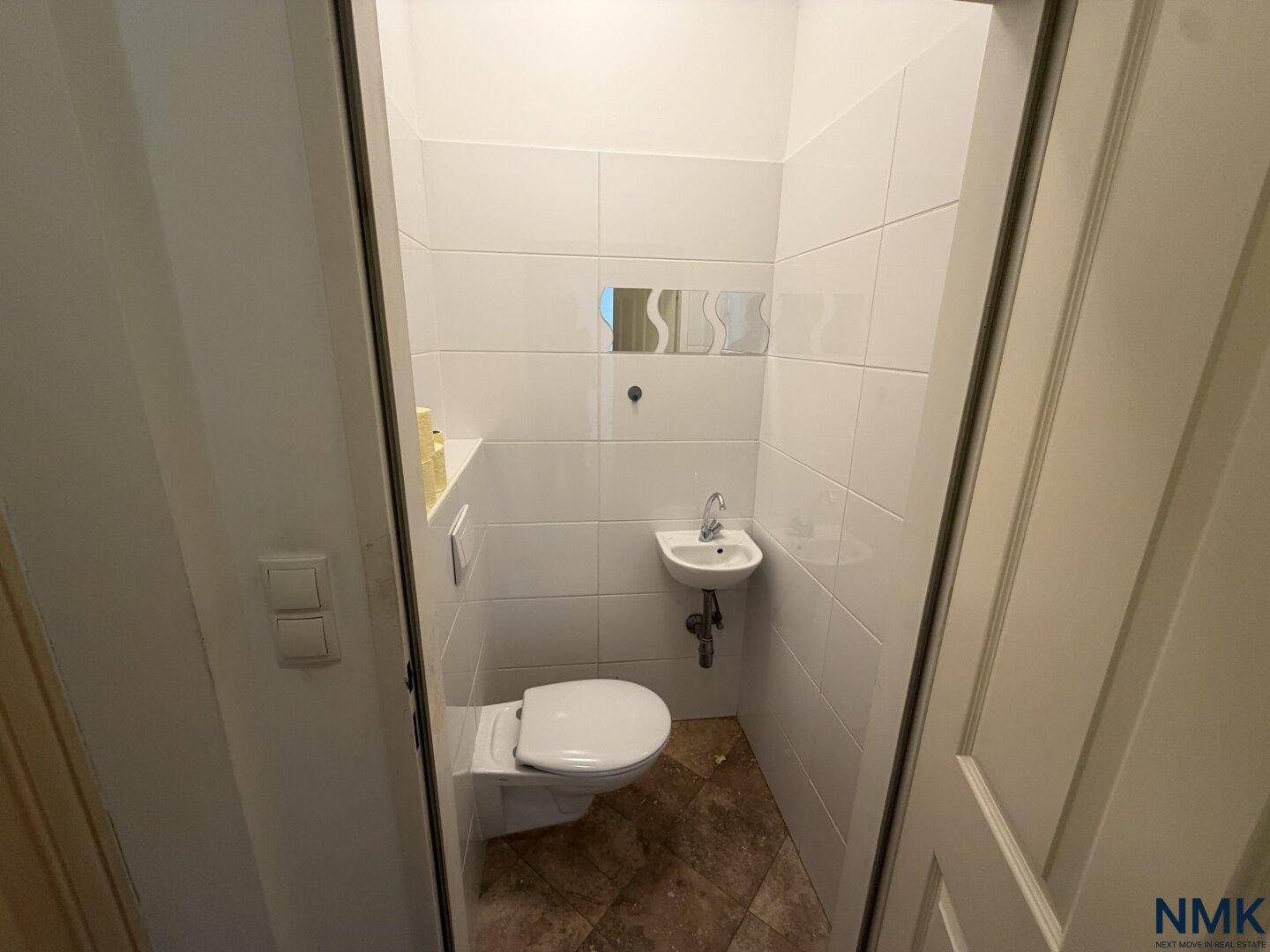 Gäste-WC im Originalzustand mit WC, kleinem Waschbecken und weißen Wandfliesen.