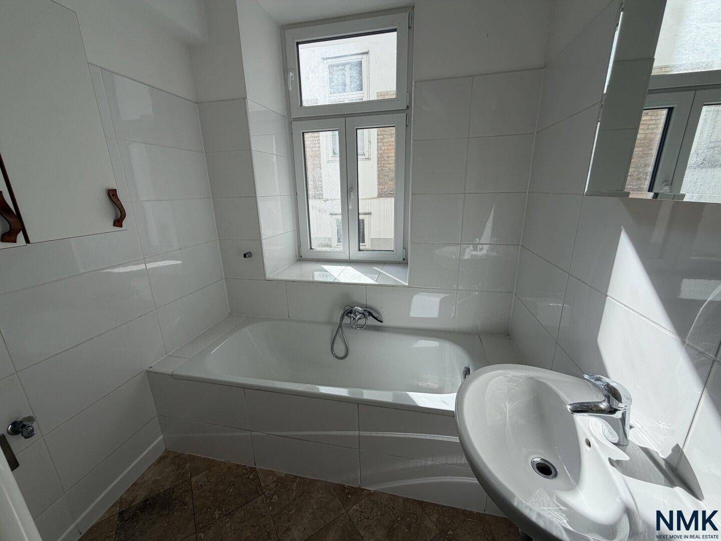 Badezimmer im Originalzustand mit Badewanne, weißen Fliesen und einem Waschbecken unter dem Fenster.