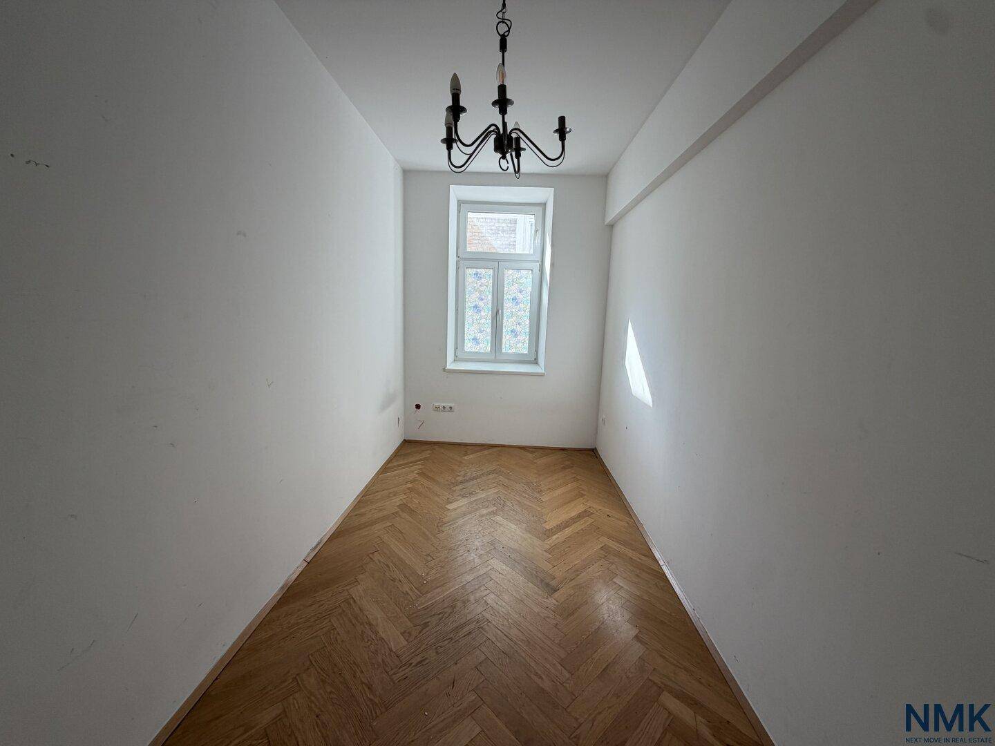Unrenovierter schmaler Raum mit Fischgrätparkett, Kronleuchter und einem Fenster im Originalzustand.