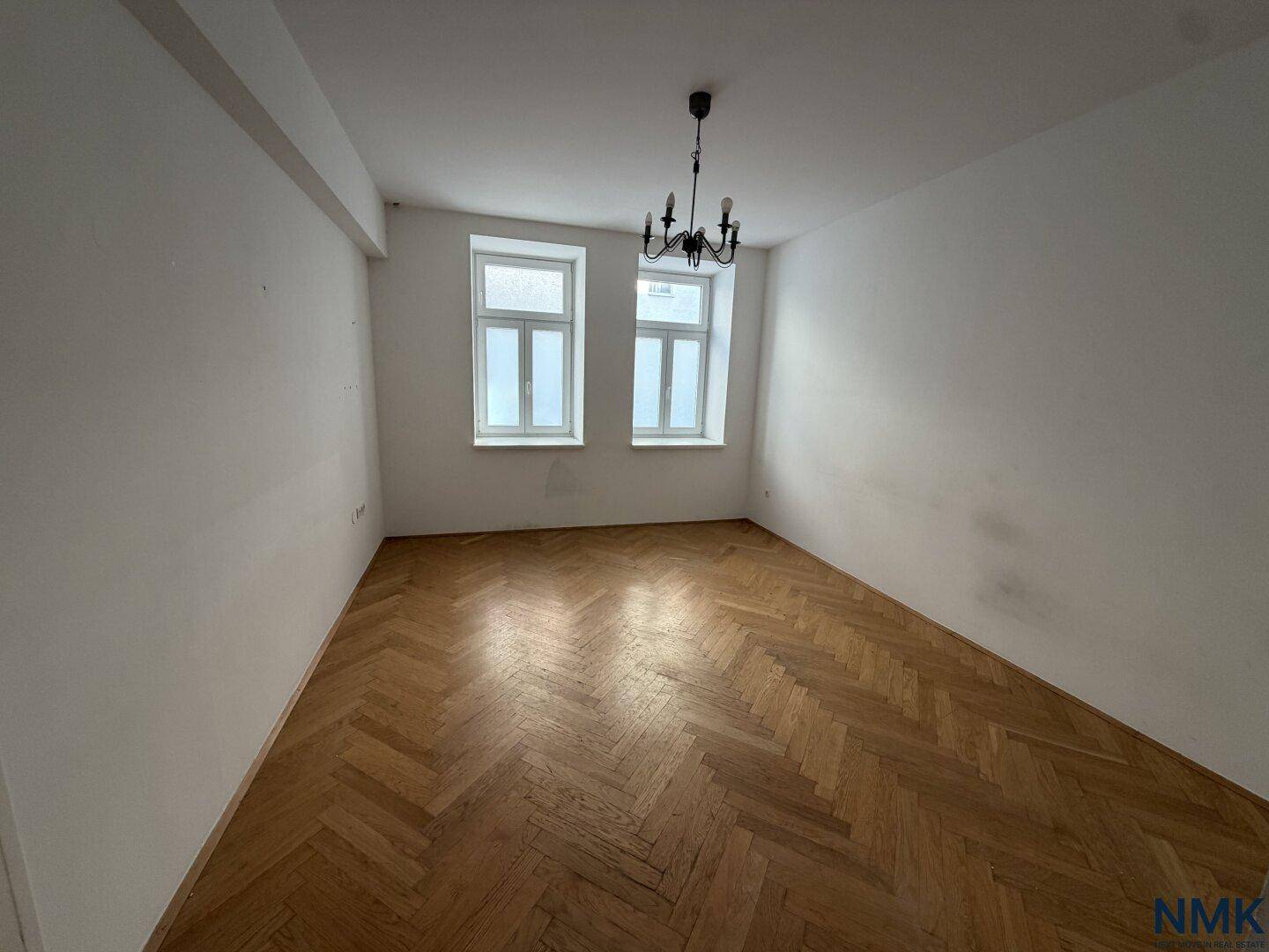 Unrenoviertes Zimmer mit Fischgrätparkett, Kronleuchter und zwei Fenstern im Originalzustand.