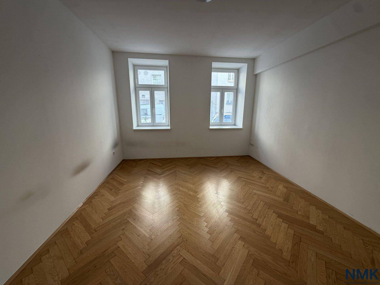 Unrenoviertes Zimmer mit Fischgrätparkett und zwei Fenstern, zeigt den Originalzustand der Immobilie.