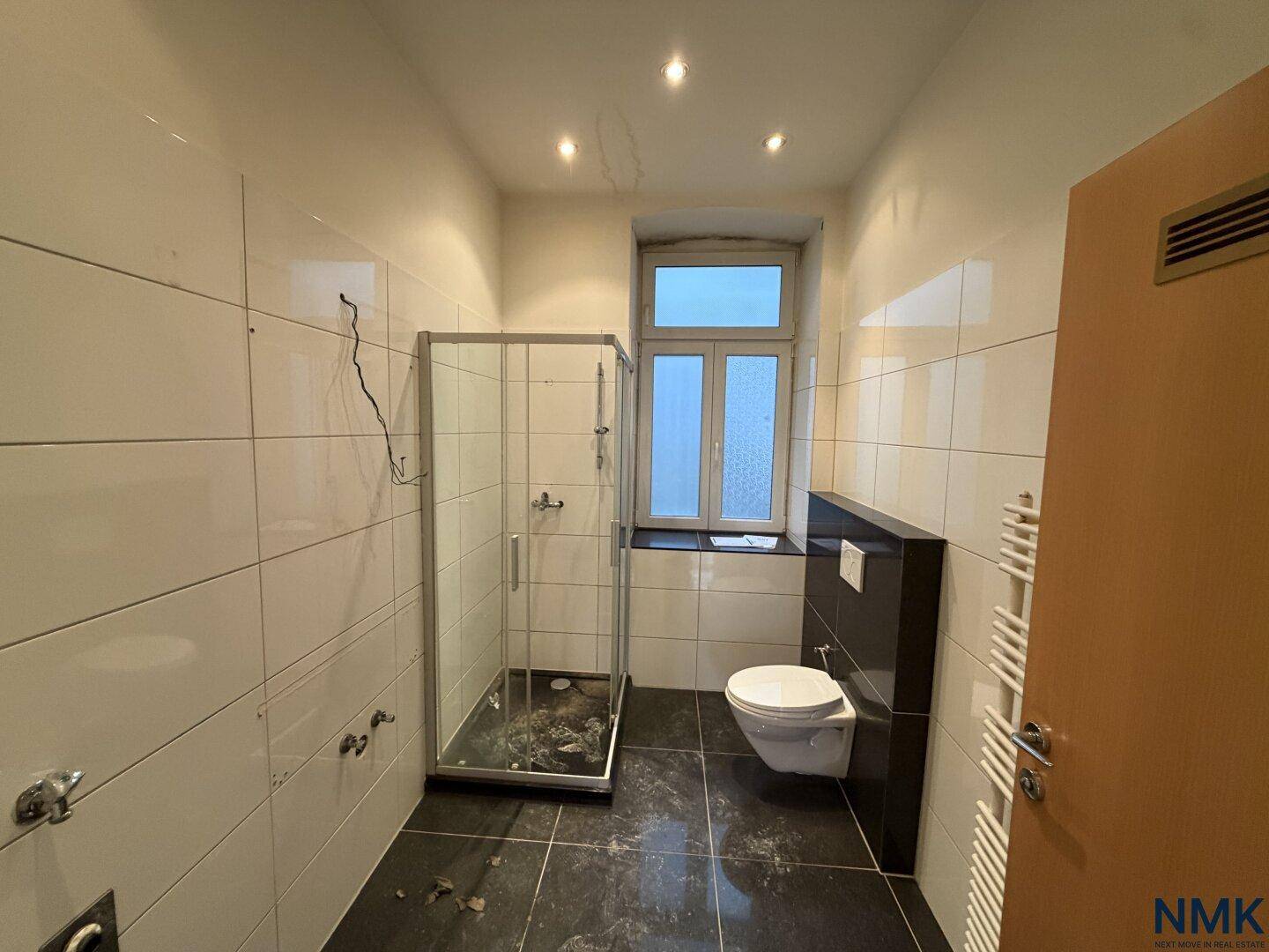 Badezimmer vor der Sanierung mit älteren Fliesen, einfacher Duschkabine und Wand-WC.