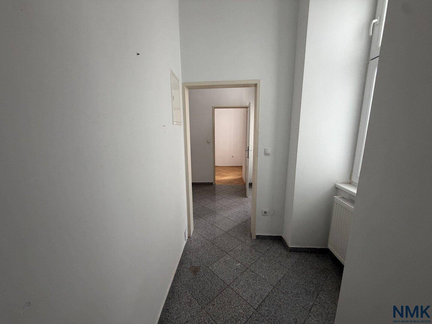Einfacher Flur mit grauen Fliesen und weißen Wänden, der Renovierungsbedarf aufweist.
