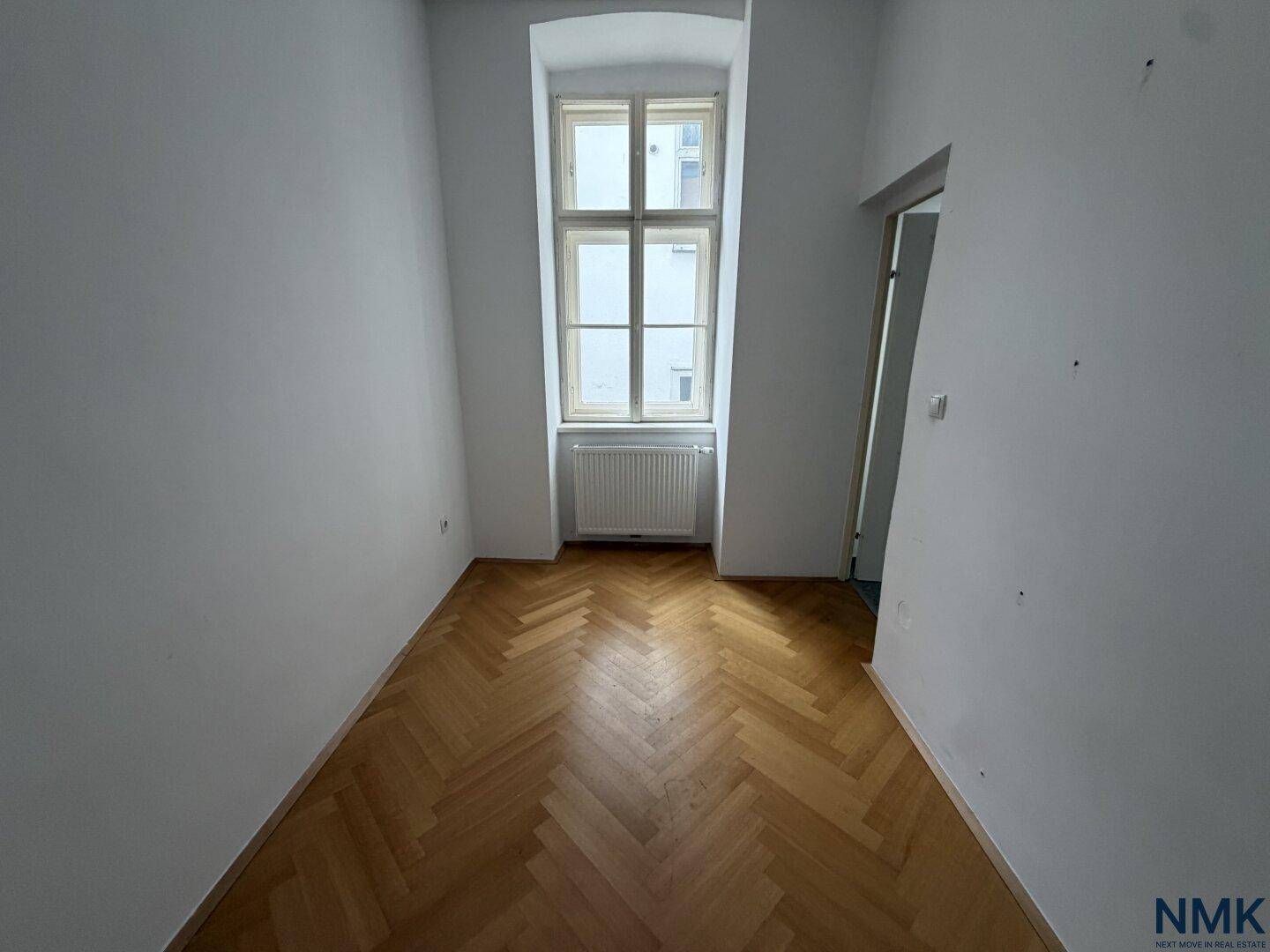 Helles Zimmer mit Fischgrätparkett und einem großen Fenster, ideal als Schlaf- oder Arbeitszimmer.