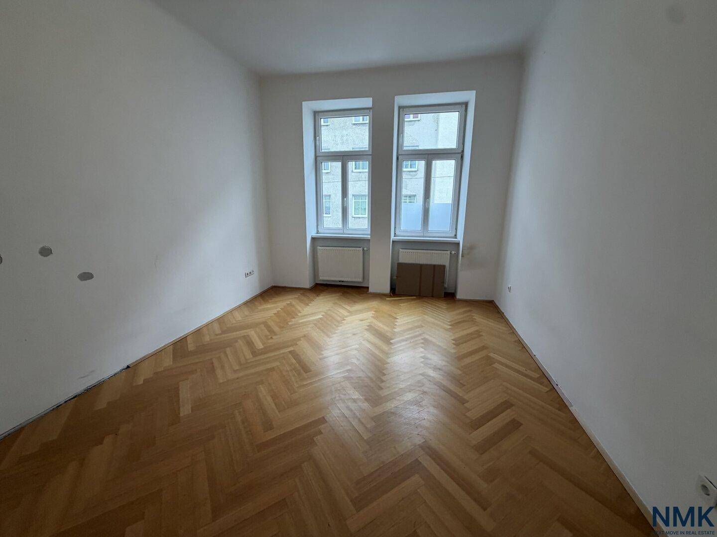 Leeres Zimmer mit klassischem Fischgrätparkett und zwei Fenstern, bereit für die individuelle Gestaltung.